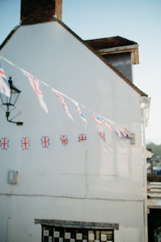 A white building with a brick chimney features a string of Union Jack flags hanging across its facade. A traditional black metal streetlamp is affixed on the side of the building near the flags. The lower part of the building shows a window with a black and white checker pattern. The sky is clear and blue, indicating daylight.