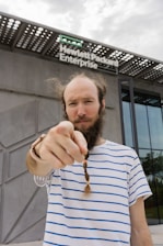 A person with a striped shirt and braided beard stands in front of a modern building with a sign that reads Hewlett Packard Enterprise. The person is pointing directly at the camera with a focused expression.
