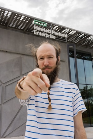 A person with a striped shirt and braided beard stands in front of a modern building with a sign that reads Hewlett Packard Enterprise. The person is pointing directly at the camera with a focused expression.