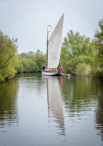 A large sailboat with a tall white sail glides down a calm river, framed by lush green trees on both sides. People stand on the boat, and a red flag is visible at the top of the mast. The reflection of the sail is visible in the water, creating a serene and picturesque scene.