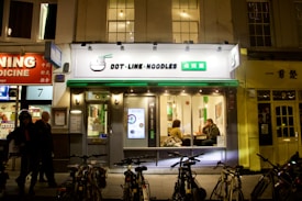 A noodle restaurant with a bright sign featuring both English and Chinese characters is situated on a busy street at night. The facade is modern with large windows through which two people can be seen sitting at a table inside. Bicycles are parked in front of the restaurant, and other businesses with illuminated signs are visible on either side.