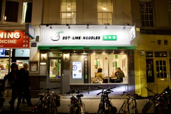 A noodle restaurant with a bright sign featuring both English and Chinese characters is situated on a busy street at night. The facade is modern with large windows through which two people can be seen sitting at a table inside. Bicycles are parked in front of the restaurant, and other businesses with illuminated signs are visible on either side.