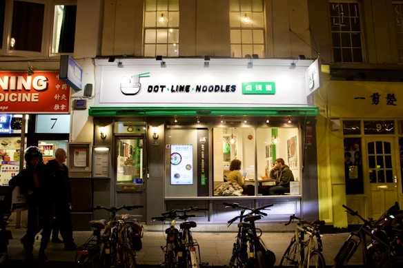 A noodle restaurant with a bright sign featuring both English and Chinese characters is situated on a busy street at night. The facade is modern with large windows through which two people can be seen sitting at a table inside. Bicycles are parked in front of the restaurant, and other businesses with illuminated signs are visible on either side.