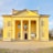 A neoclassical building with a bright yellow facade featuring four tall white columns at the entrance. The structure includes several arched windows with shutters and a large wooden door in the center. Flags are displayed above the door, and there is a bulletin board attached to the front wall. The building is set against a clear blue sky and surrounded by trees.