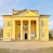 A neoclassical building with a bright yellow facade featuring four tall white columns at the entrance. The structure includes several arched windows with shutters and a large wooden door in the center. Flags are displayed above the door, and there is a bulletin board attached to the front wall. The building is set against a clear blue sky and surrounded by trees.