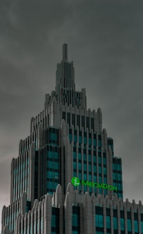 A tall, modern skyscraper with a detailed, tiered architectural design standing against a cloudy sky. The building features multiple levels with geometric precision and a distinctive spire at the top. A green sign with the text in Cyrillic script is prominently displayed near the upper section.