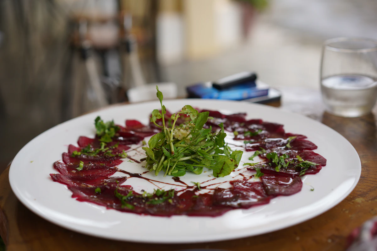 A fine-dining beetroot carpaccio fanned on a white plate with watercress, herbs and balsamic.