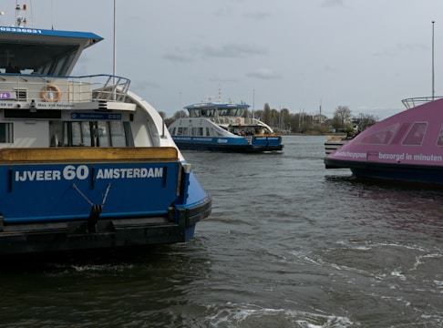 Three ferries are on a body of water, with the closest one displaying 'IJVEER 60 AMSTERDAM' on its stern. The middle ferry is positioned in the distance, partially obstructed, and the ferry on the right has a pink exterior.
