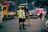 A police officer is directing traffic on a busy street. He is wearing a reflective vest and holding a baton. A rickshaw with a passenger is nearby, while several vehicles, including a yellow truck and a sedan with red lights, are visible on the road.
