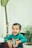 A young child with short hair is sitting indoors wearing a green shirt with a colorful design. The child is smiling joyfully. In the background, there are green plants, and the room has a bright and fresh atmosphere.
