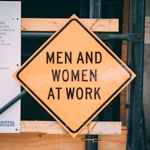A construction warning sign in the shape of a diamond with the words 'Men and Women at Work' is prominently displayed. The sign is yellow with black text and is set against a background of construction materials including wood planks and metal scaffolding.