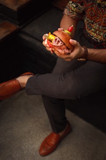 A person is holding a gourmet sandwich filled with layers of sliced meat, pickles, and garnished with a small skewer of red cherry tomatoes and a pepper. The person is seated, wearing patterned clothing and brown loafers, and has one leg crossed over the other.