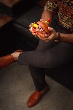 A person is holding a gourmet sandwich filled with layers of sliced meat, pickles, and garnished with a small skewer of red cherry tomatoes and a pepper. The person is seated, wearing patterned clothing and brown loafers, and has one leg crossed over the other.