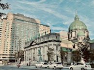A large hotel building with many windows is situated next to a historic cathedral with a domed roof and statues adorning its exterior. Several cars are visible on the street in the foreground. Trees line the sidewalk, and a Canadian flag waves atop the building.