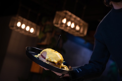 A person holding a black plate with a sandwich topped with a generous amount of melted yellow cheese. The background features hanging lights, creating a warm and dimly-lit atmosphere likely set in a restaurant or cafe.