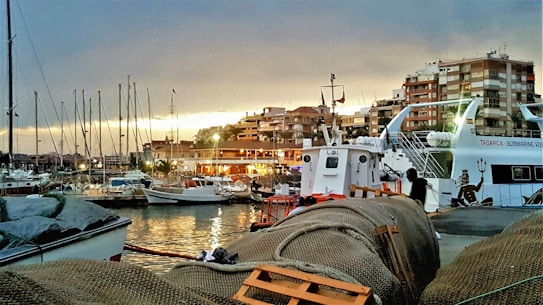 A harbor scene at dusk with various sailing boats docked along the water. Buildings with orange and brown tones are visible in the background, illuminated with warm lights. Fishing nets and equipment are prominently placed in the foreground, and a large boat is situated to the right, displaying the words 'Tabarca Submarine Vision' along with graphics.