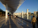 A modern architectural structure with white geometric columns and a wavy roof extends along a waterfront promenade. People walk and cycle along the tiled pathway, casting long shadows in the sunlight. The background features a city skyline with several high-rise buildings and a clear blue sky.
