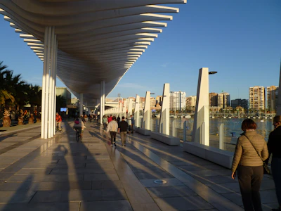 A modern architectural structure with white geometric columns and a wavy roof extends along a waterfront promenade. People walk and cycle along the tiled pathway, casting long shadows in the sunlight. The background features a city skyline with several high-rise buildings and a clear blue sky.