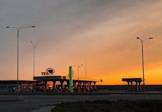 A gas station is illuminated by the warm, orange glow of a sunset, casting shadows on the ground. Tall street lamps stand on either side, silhouetted against the sky. The background shows a flat landscape with a few scattered buildings.