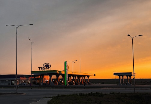 A gas station is illuminated by the warm, orange glow of a sunset, casting shadows on the ground. Tall street lamps stand on either side, silhouetted against the sky. The background shows a flat landscape with a few scattered buildings.
