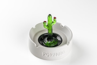 A glass sculpture resembling a green cactus stands at the center of a white ceramic ashtray. The sculpture features two red tips on the cactus arms, adding a touch of color contrast. The ashtray has the word 'PORFIDIO' engraved on its side.