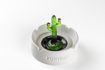 A glass sculpture resembling a green cactus stands at the center of a white ceramic ashtray. The sculpture features two red tips on the cactus arms, adding a touch of color contrast. The ashtray has the word 'PORFIDIO' engraved on its side.