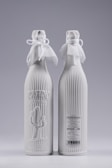 Two elegant white bottles with ribbed texture and decorative cloth tied around the neck with bows. One bottle has embossed text and a cactus design on the front, while the other displays product details and a barcode.