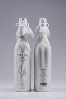 Two elegant white bottles with ribbed texture and decorative cloth tied around the neck with bows. One bottle has embossed text and a cactus design on the front, while the other displays product details and a barcode.