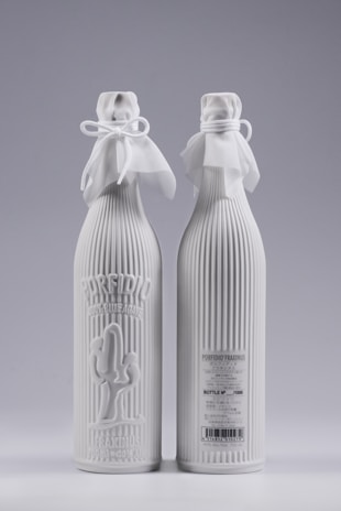 Two elegant white bottles with ribbed texture and decorative cloth tied around the neck with bows. One bottle has embossed text and a cactus design on the front, while the other displays product details and a barcode.
