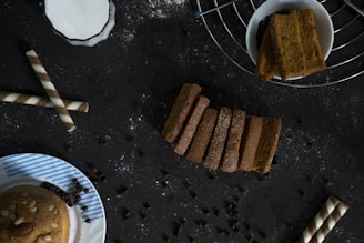 A dark-toned background features an assortment of baked goods and snacks. In the center, several slices of bread are neatly arranged, sprinkled with some white powder. To the right, a bowl contains thick, rectangular slices, positioned on a wire rack. To the left, two wafer rolls are placed diagonally, and a partly visible plate with a muffin topped with almonds and seeds sits at the bottom left corner. Scattered chocolate chips are spread across the scene.