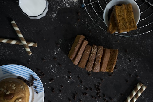A dark-toned background features an assortment of baked goods and snacks. In the center, several slices of bread are neatly arranged, sprinkled with some white powder. To the right, a bowl contains thick, rectangular slices, positioned on a wire rack. To the left, two wafer rolls are placed diagonally, and a partly visible plate with a muffin topped with almonds and seeds sits at the bottom left corner. Scattered chocolate chips are spread across the scene.
