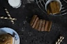 A dark-toned background features an assortment of baked goods and snacks. In the center, several slices of bread are neatly arranged, sprinkled with some white powder. To the right, a bowl contains thick, rectangular slices, positioned on a wire rack. To the left, two wafer rolls are placed diagonally, and a partly visible plate with a muffin topped with almonds and seeds sits at the bottom left corner. Scattered chocolate chips are spread across the scene.
