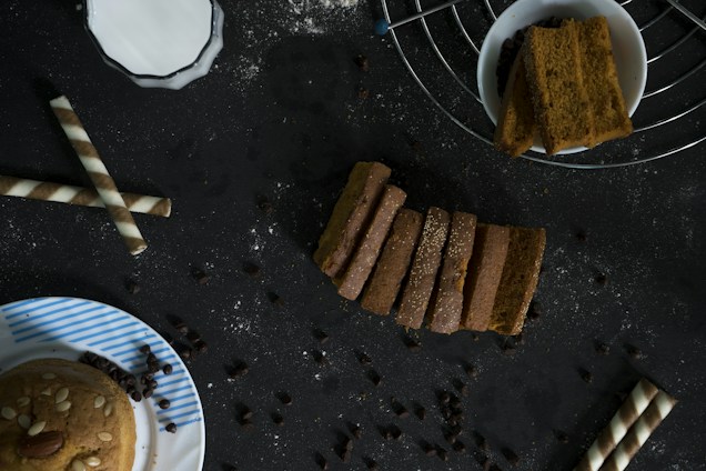 A dark-toned background features an assortment of baked goods and snacks. In the center, several slices of bread are neatly arranged, sprinkled with some white powder. To the right, a bowl contains thick, rectangular slices, positioned on a wire rack. To the left, two wafer rolls are placed diagonally, and a partly visible plate with a muffin topped with almonds and seeds sits at the bottom left corner. Scattered chocolate chips are spread across the scene.