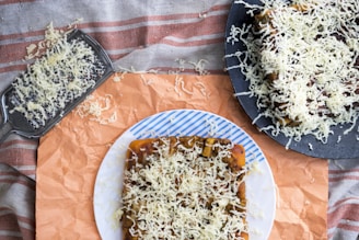 A piece of bread topped with shredded cheese is displayed on a white plate with blue stripes. Additional bread with cheese and a metal cheese grater are visible, set on a patterned cloth and orange paper.