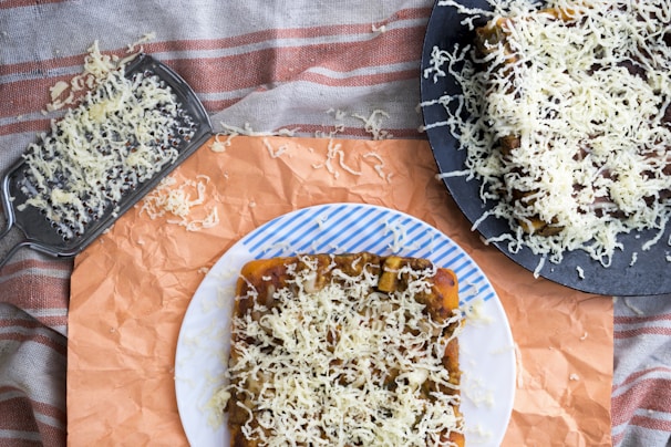 A piece of bread topped with shredded cheese is displayed on a white plate with blue stripes. Additional bread with cheese and a metal cheese grater are visible, set on a patterned cloth and orange paper.