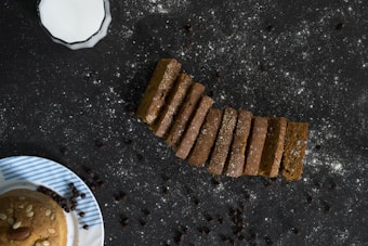 A set of brown bread slices arranged in a neat line on a dark surface sprinkled with flour and chocolate chips. In the top left corner, there's a cup of milk with a decorative edge. At the bottom, part of a striped ceramic plate with a round pastry topped with nuts is visible.
