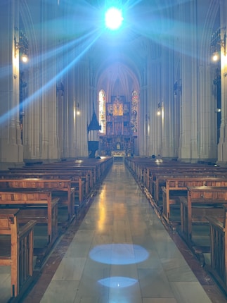 An expansive, ornate cathedral interior with high vaulted ceilings, intricately designed stained glass windows, and wooden pews lining both sides of a long central aisle. A bright, radiant light shines from above, casting blue-tinted illumination across the scene.