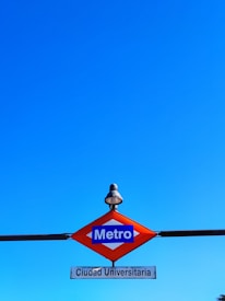 A metro sign depicting the station 'Ciudad Universitaria' is displayed against a clear blue sky. The sign has a distinct diamond shape with a red border and a blue center where 'Metro' is written in white letters. Below the diamond, there's a white rectangular plate with the name 'Ciudad Universitaria' in black lettering. Above the sign, a small, vintage-style streetlamp is present.