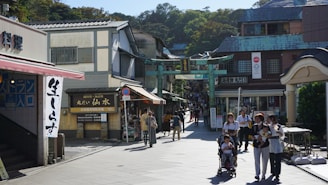 A busy street scene in an Asian town featuring a market area with traditional architecture. Several people, including a family with a stroller, are walking by, suggesting a bustling and lively atmosphere. Shops line the street with signs featuring Asian script.