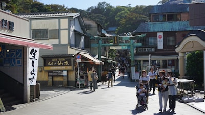 A busy street scene in an Asian town featuring a market area with traditional architecture. Several people, including a family with a stroller, are walking by, suggesting a bustling and lively atmosphere. Shops line the street with signs featuring Asian script.