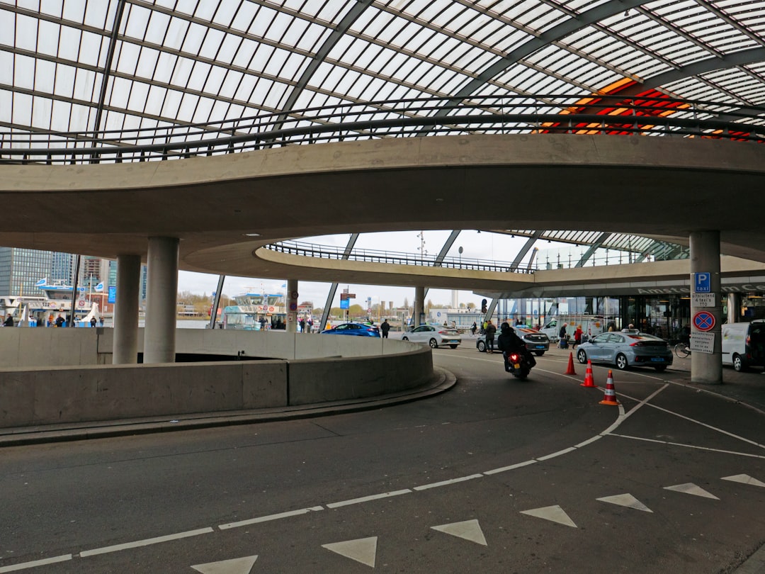 a road with cars and a bridge, A view on the west side of the bus station, just behind train station Amsterdam. It is rather spatial modern architecture here with a light view over river IJ and the boats. Free urban photo of Central station building Amsterdam, Fons Heijnsbroek - The Netherlands, 30 March 2022 // Gratis foto, moderne station architectuur & design, Amsterdam - gratis downloaden: het busstation met dak en westelijke oprijbaan, achter Centraal Station - zomer 2022. Foto, Fons Heijnsbroek - stations-fotografie, Nederland in hoge resolutie.