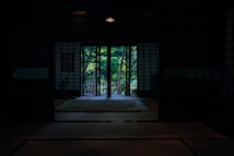 A dimly lit room with traditional Japanese architecture featuring shoji screens and tatami mats. A view through the open sliding doors reveals lush greenery and a well-kept garden outside. A soft carpet is placed in the center of the room, and a decorative scroll or painting hangs on the wall.