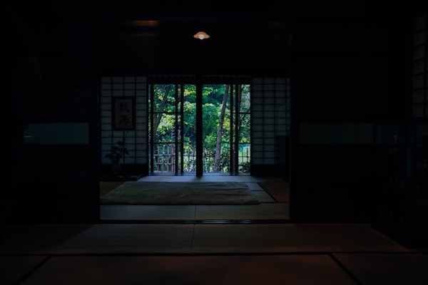 A dimly lit room with traditional Japanese architecture featuring shoji screens and tatami mats. A view through the open sliding doors reveals lush greenery and a well-kept garden outside. A soft carpet is placed in the center of the room, and a decorative scroll or painting hangs on the wall.
