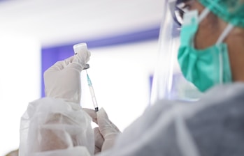 A healthcare professional wearing protective attire, including a face mask and shield, is preparing a syringe with a vaccine from a small vial. The setting appears to be clinical with an emphasis on safety and hygiene.