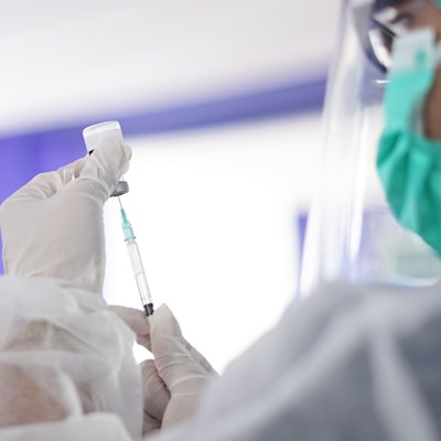 A healthcare professional wearing protective attire, including a face mask and shield, is preparing a syringe with a vaccine from a small vial. The setting appears to be clinical with an emphasis on safety and hygiene.