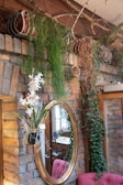 A rustic interior setting featuring a brick wall adorned with various green plants. Hanging baskets contain cascading foliage, including ferns and ivy. A round, gold-framed mirror reflects part of the room, showing a hint of furniture and decor. Below the mirror, there is a vase with white flowers. The overall ambiance is cozy and lush.