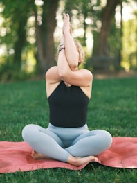 A person sits cross-legged on a yoga mat in a grassy outdoor setting, performing a yoga pose with arms intertwined in front of their face. The trees and foliage in the background provide a natural, serene environment.