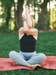 A person sits cross-legged on a yoga mat in a grassy outdoor setting, performing a yoga pose with arms intertwined in front of their face. The trees and foliage in the background provide a natural, serene environment.