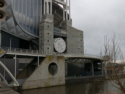 The image depicts a large, modern architectural structure with a prominent concrete facade and circular openings. A logo with the text 'Ajax' and an illustration is visible on the building. There are metal railings and a bridge-like walkway, and a bare tree is visible in the foreground. The sky is overcast, suggesting a cloudy day.