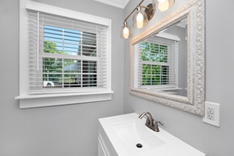 A bathroom interior with a large ornate mirror reflecting a window with white blinds. The window is open to a view of green trees outside. The wall color is light gray, and the sink has a silver faucet under three overhead lights with glass covers.